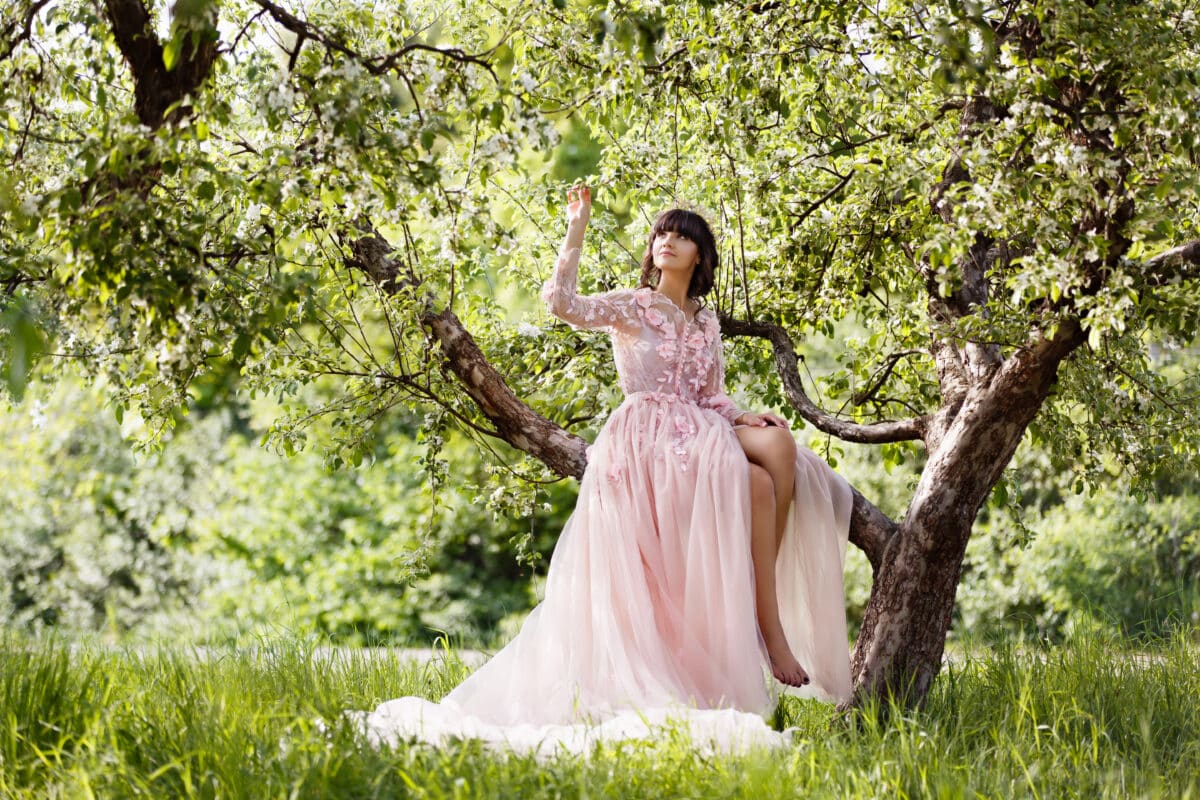 Beautiful woman in flying airy pink dress sit on a branch of blooming apple tree. Pretty young Caucasian girl plays with her dress. Girl dance in flowing dress. Romantic scene in spring garden