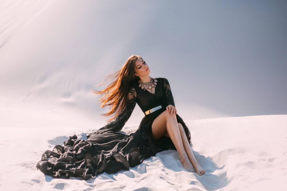Mysterious woman in a black long dress sits in desert. Luxurious clothes, gold accessories, pretty face. Oriental fashion model. Sand dunes background. Art photo.