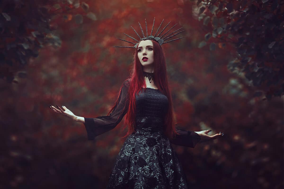 A beautiful woman with pale skin and long red hair in a black dress and in the black crownk. Girl witch with vampire claws and red lips. Gothic look. Outfit for halloween.