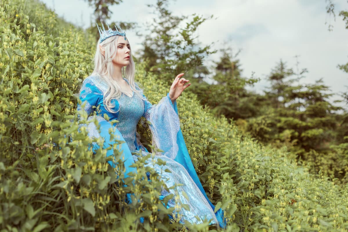 Beautiful elf woman in a blue dress is sitting in the grass on a hill.