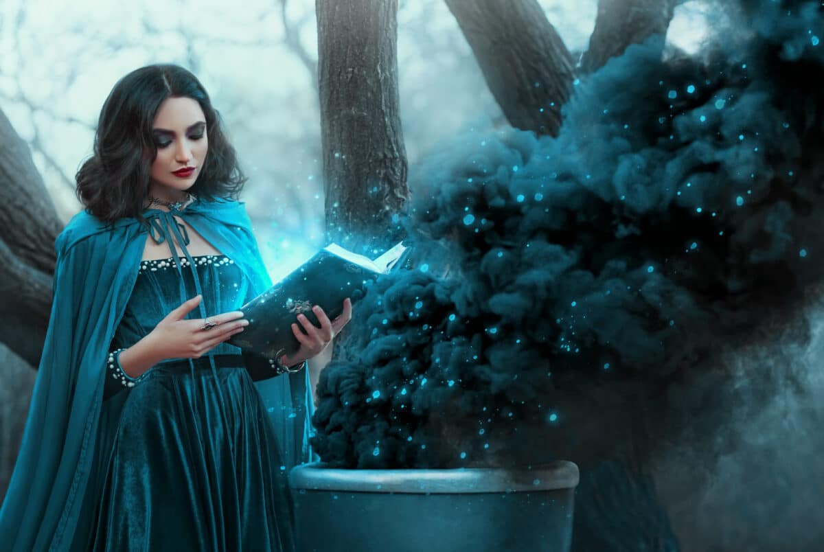 Halloween woman witch conjures, holds book in hands reads spell black magic smoke rises from boiling vat. Gothic sexy girl magician sorceress. Blue medieval dress cape. Forest trees dark autumn nature