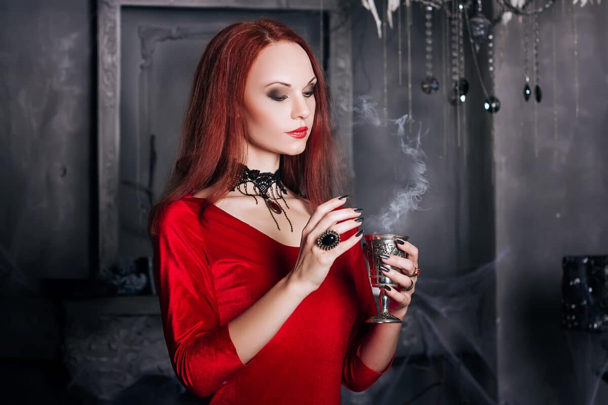 Beautiful red-haired girl with red lips. A model in a red dress with a black necklace in the castle. Vampire style. The image for halloween. Fantasy. Iron cup with wine