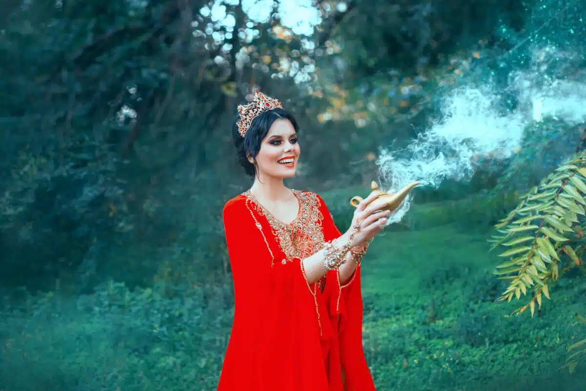 A woman in red luxurious oriental with a golden Aladdin lamp calls Gin. Positive emotions, the queen smiles. Magic and miracle. Smoke from a jug. Background summer and green trees. Art photography