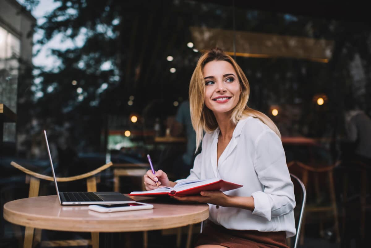 Smiling freelancer working on laptop and writing notes on terrace of cafe