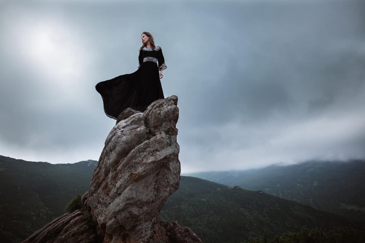 Young woman in long dress enjoying nature on the mountains. The girl in black dress with long flying train stands on the top of the rock