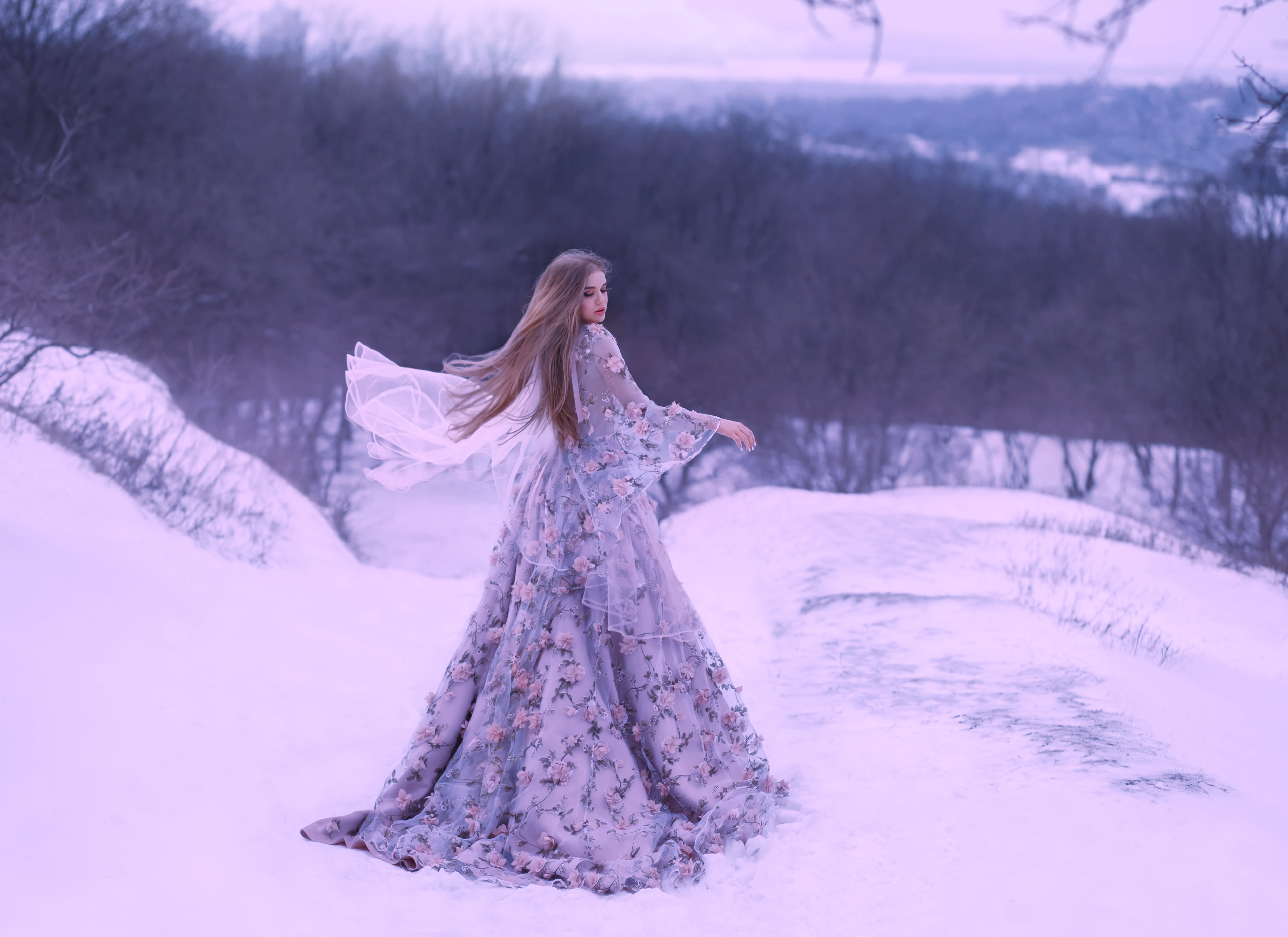 a young girl with straight fair hair in a light flying flying in the wind a purple lilac long dress decorated with flowers stands on a hill in winter on snow in the forest. art photo in cold colors.