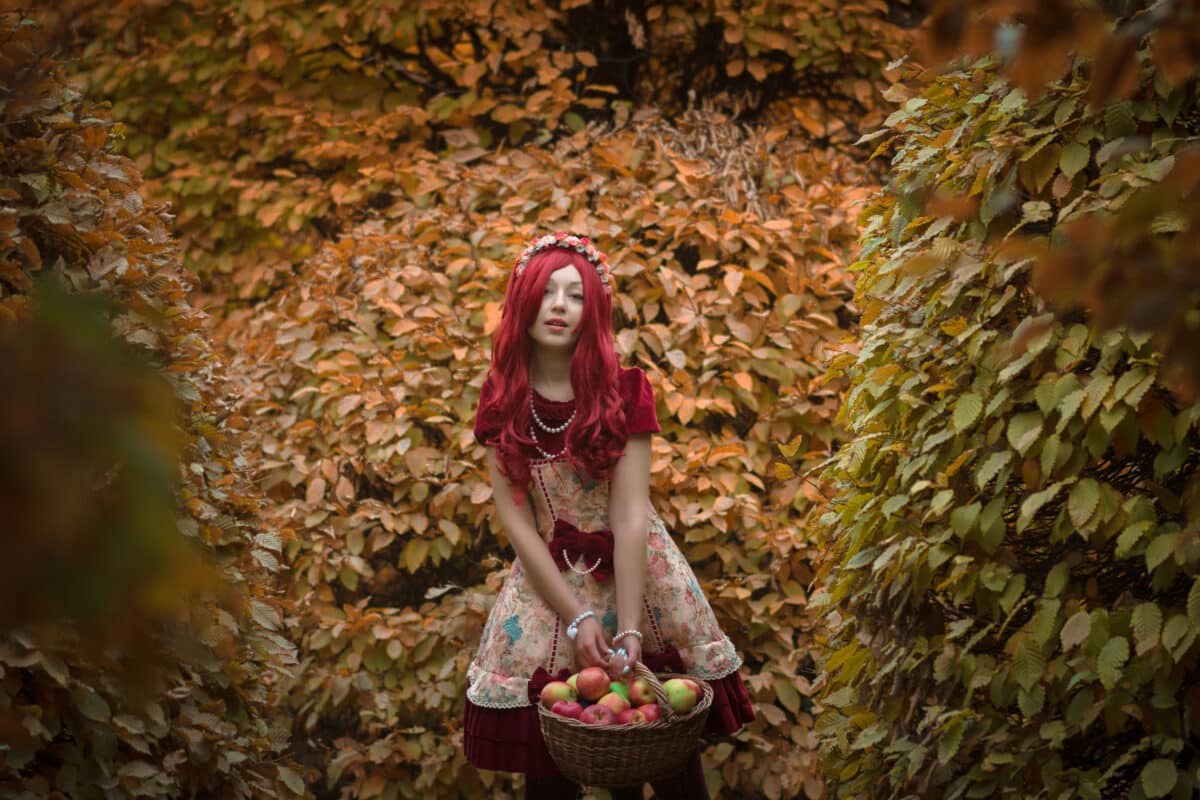 redhead woman in long dress holds heavy basket with apples in autumn forest