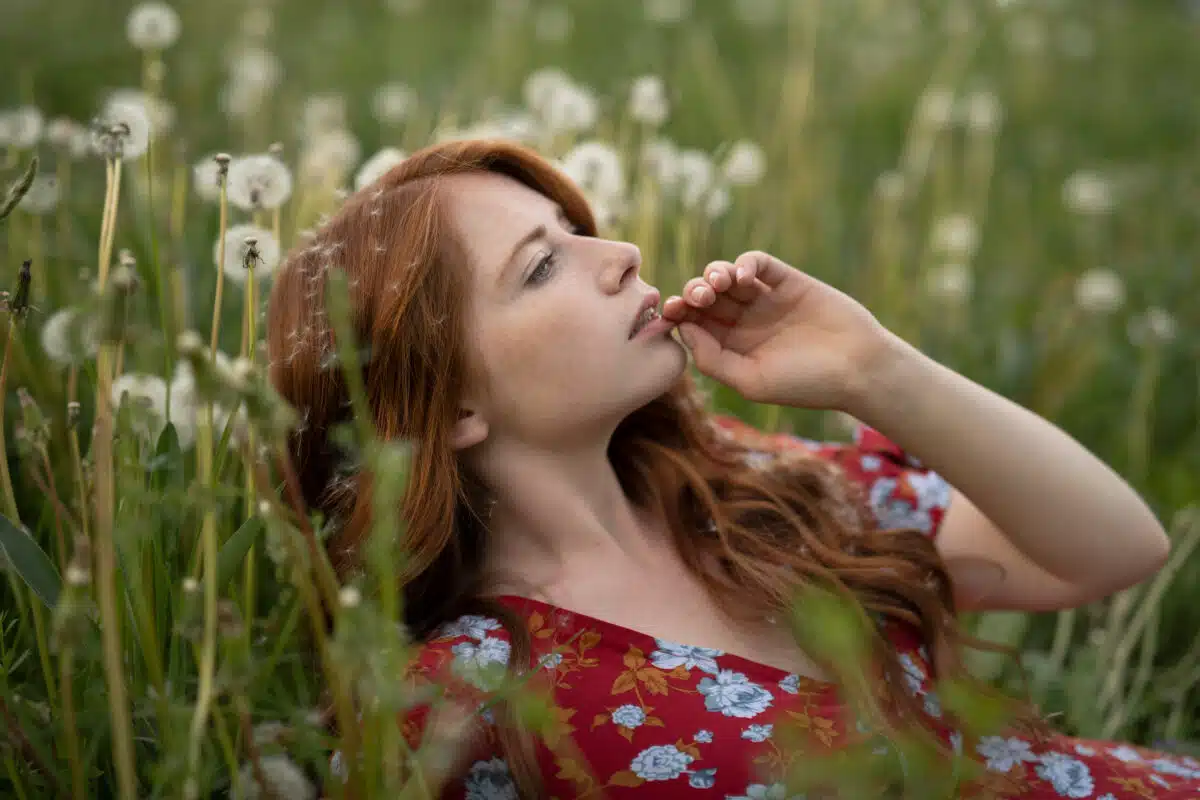 Red-haired girl in a red dress in a field among fading dandelion