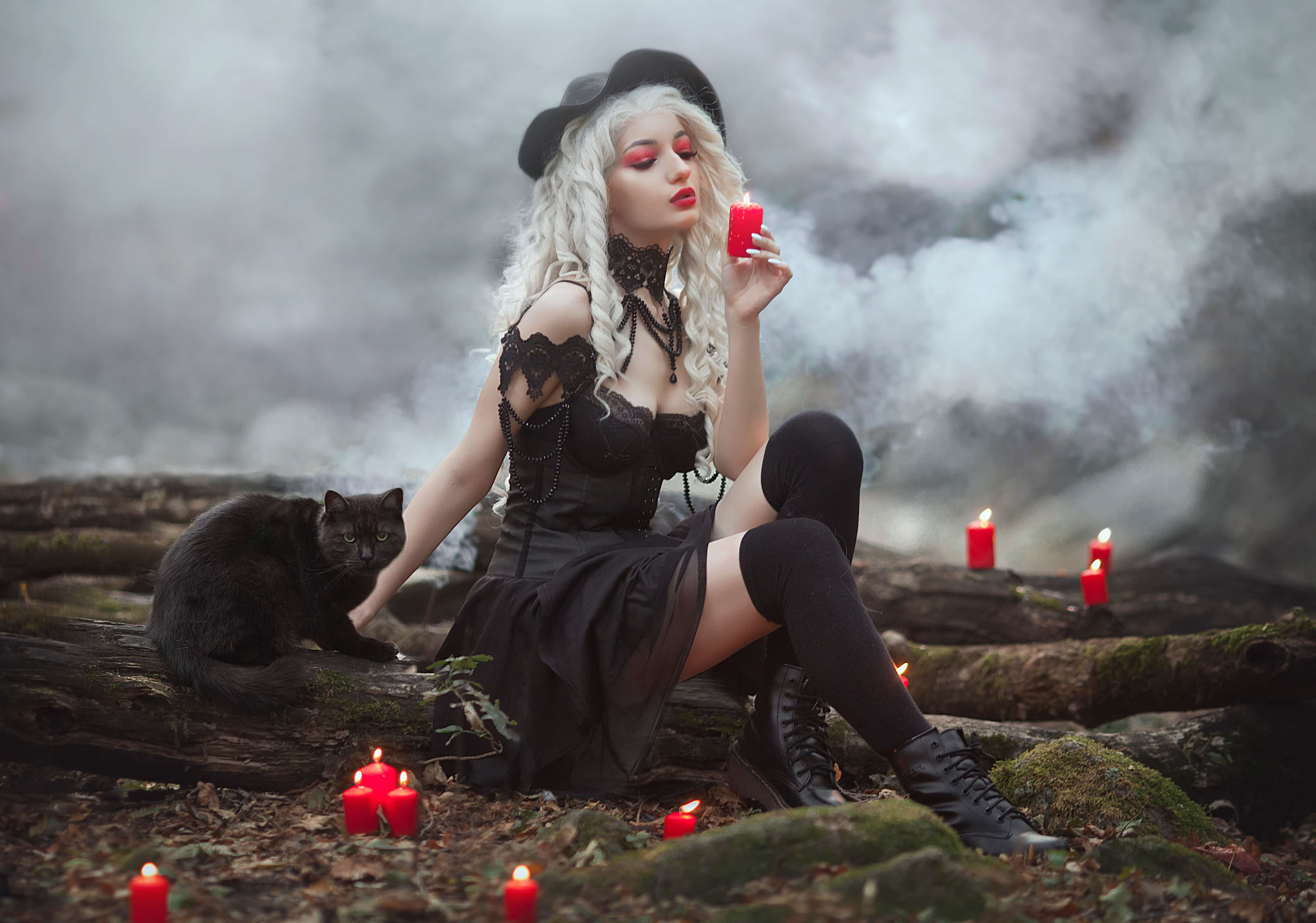Witch girl in a hat with a black cat in smoke holds red candles. Sexy witch in the forest.