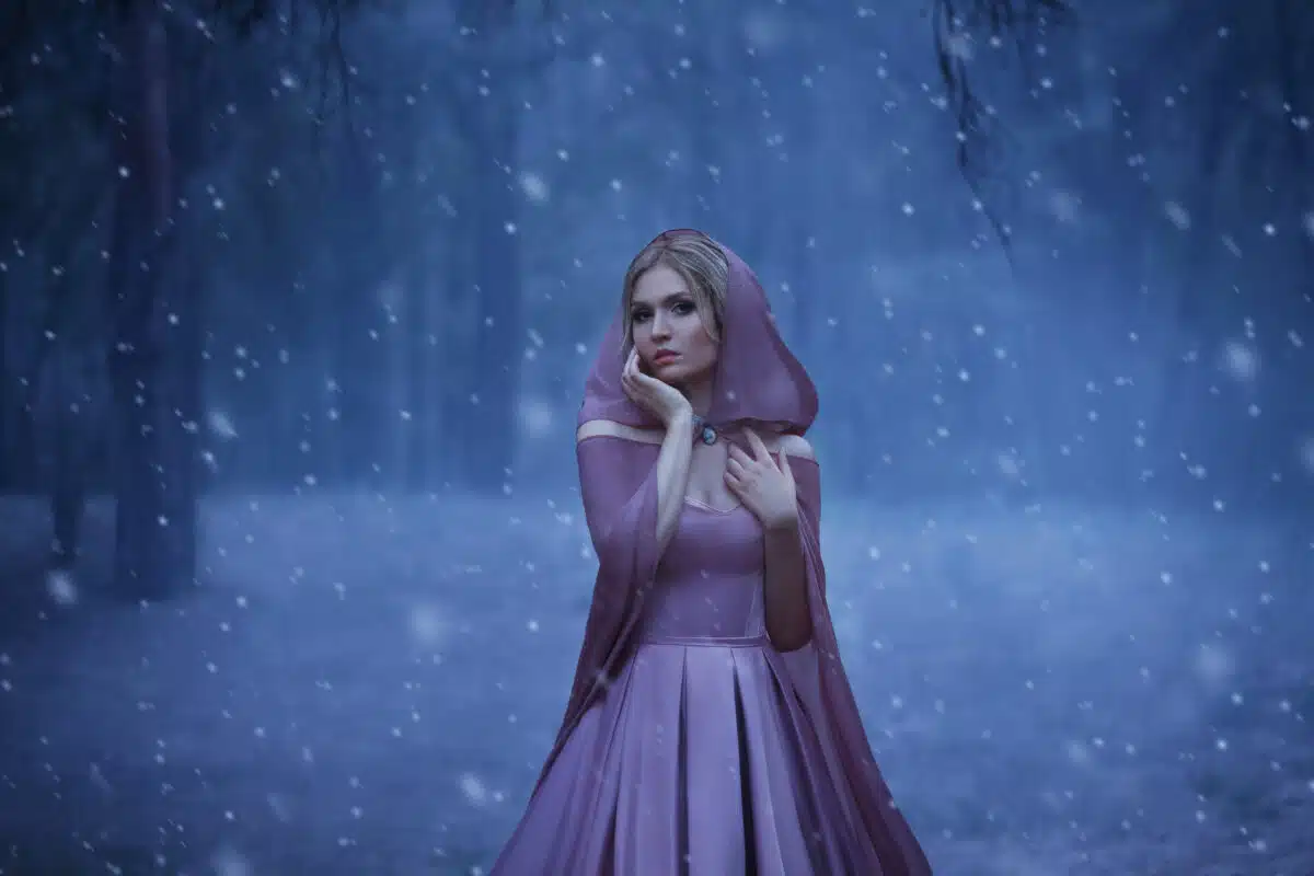 The blonde girl, a bright elf, walks in a gloomy forest covered with fog. It's cold, the snow breaks. The princess in a purple dress and in a cloak with a hood. Art photo in blue shades
