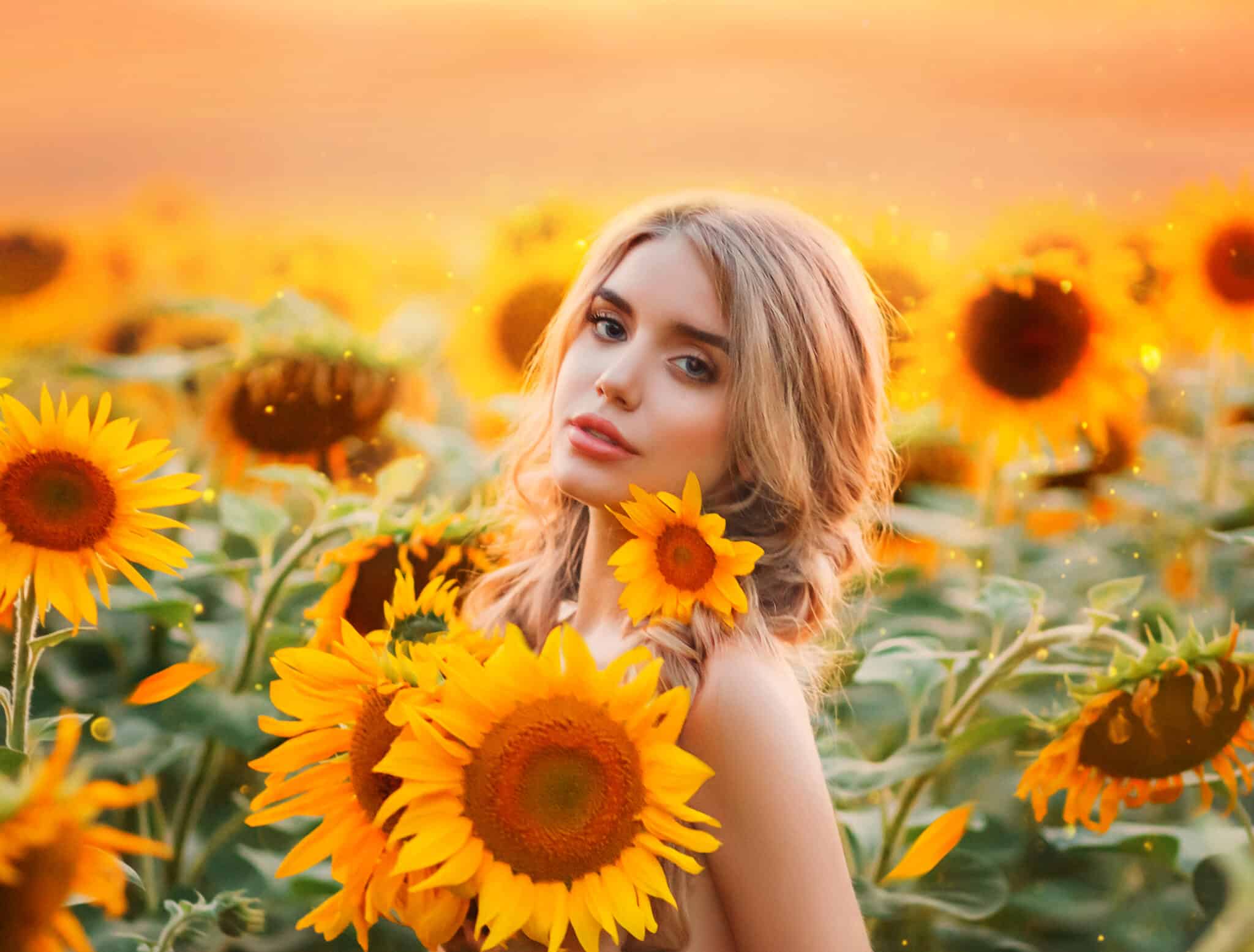 A young blonde woman stands in a field with blooming sunflowers and hugs a bouquet of flowers.