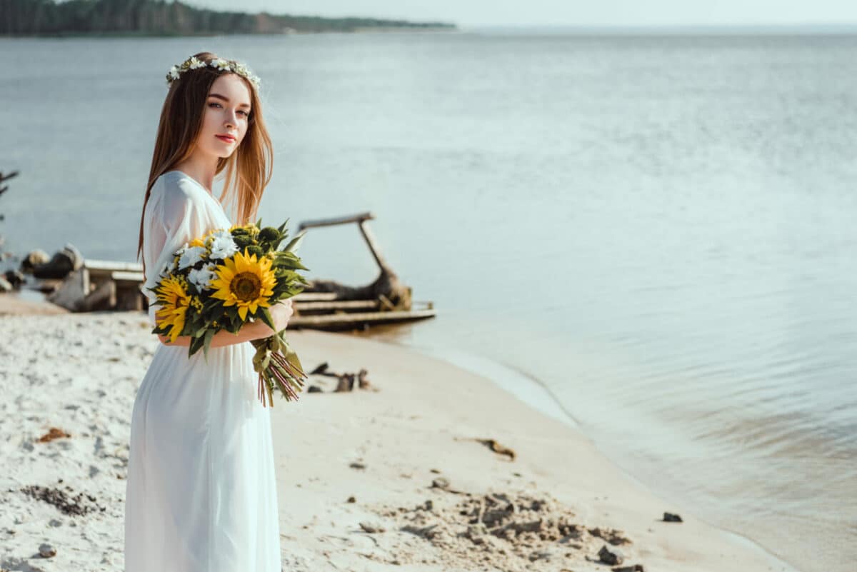 beautiful girl in white dress standing on the beach with floral head wreath holding a bouquet of sunflowers