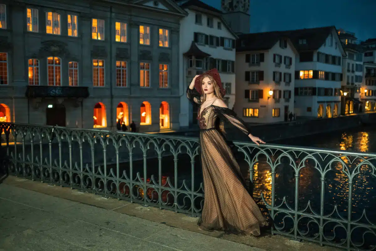 A stylish bride in a black wedding dress and a red hat poses at night in the old town of Zurich. Switzerland