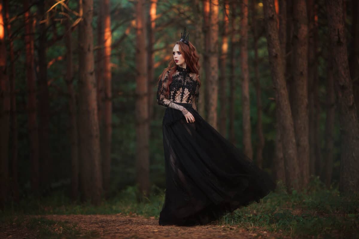 A beautiful gothic princess with pale skin and very long red hair in a black crown and a black long dress in a misty fairy forest. The costume of the dark queen.