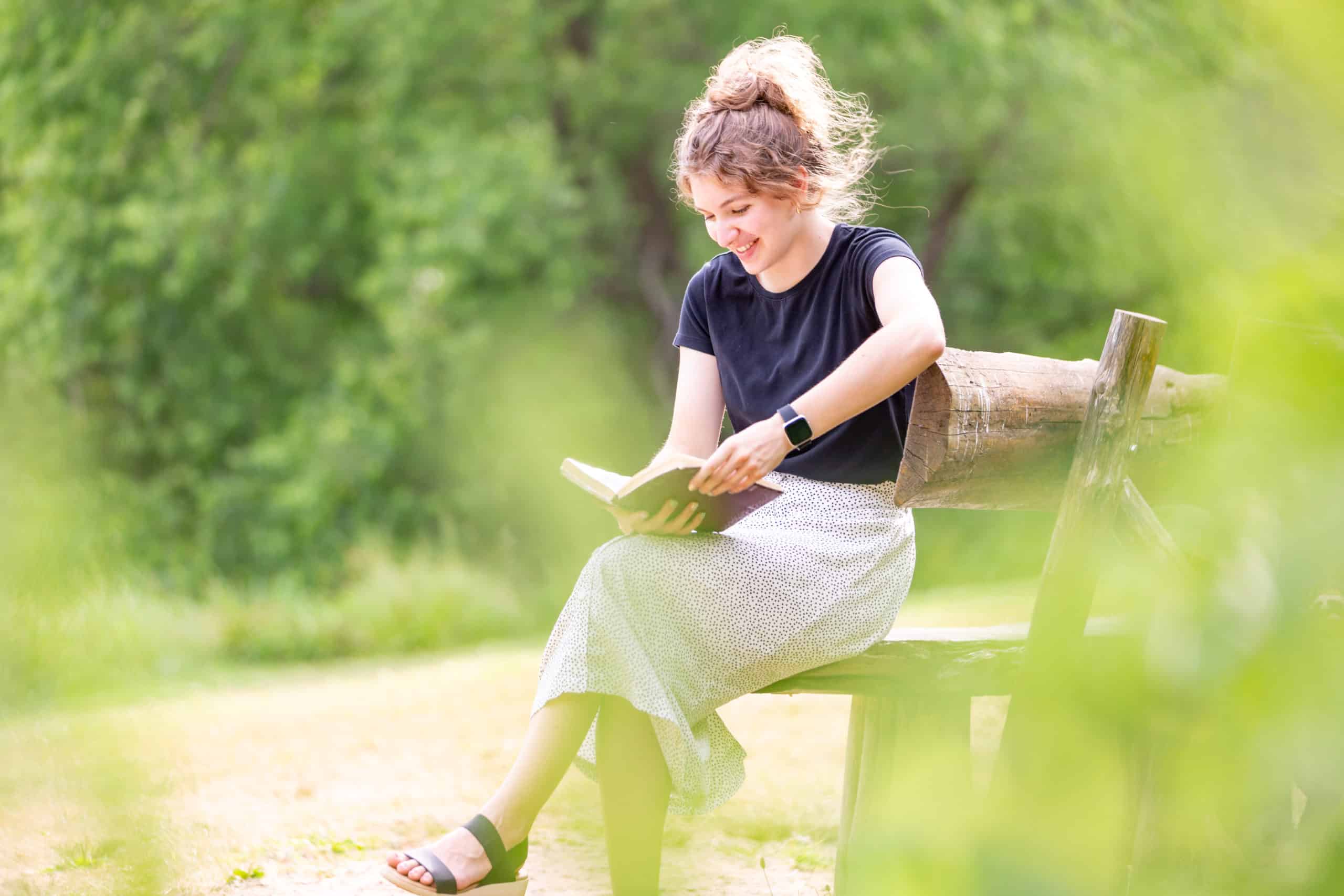 Woman smiling as she reads a book on a bench.