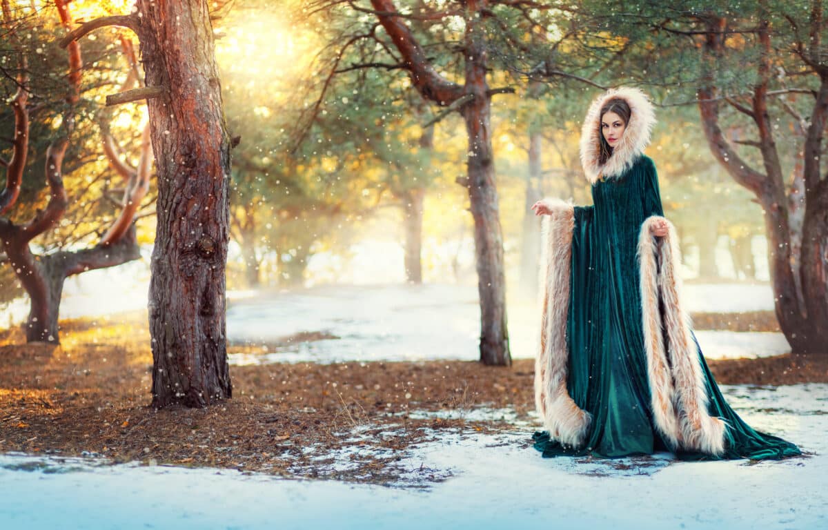 beautiful medieval woman walks in winter forest. Lady sorceress, princess girl in green long velvet vintage coat, hood with fur on head. Nature background pine tree, falling snow, magical sun light