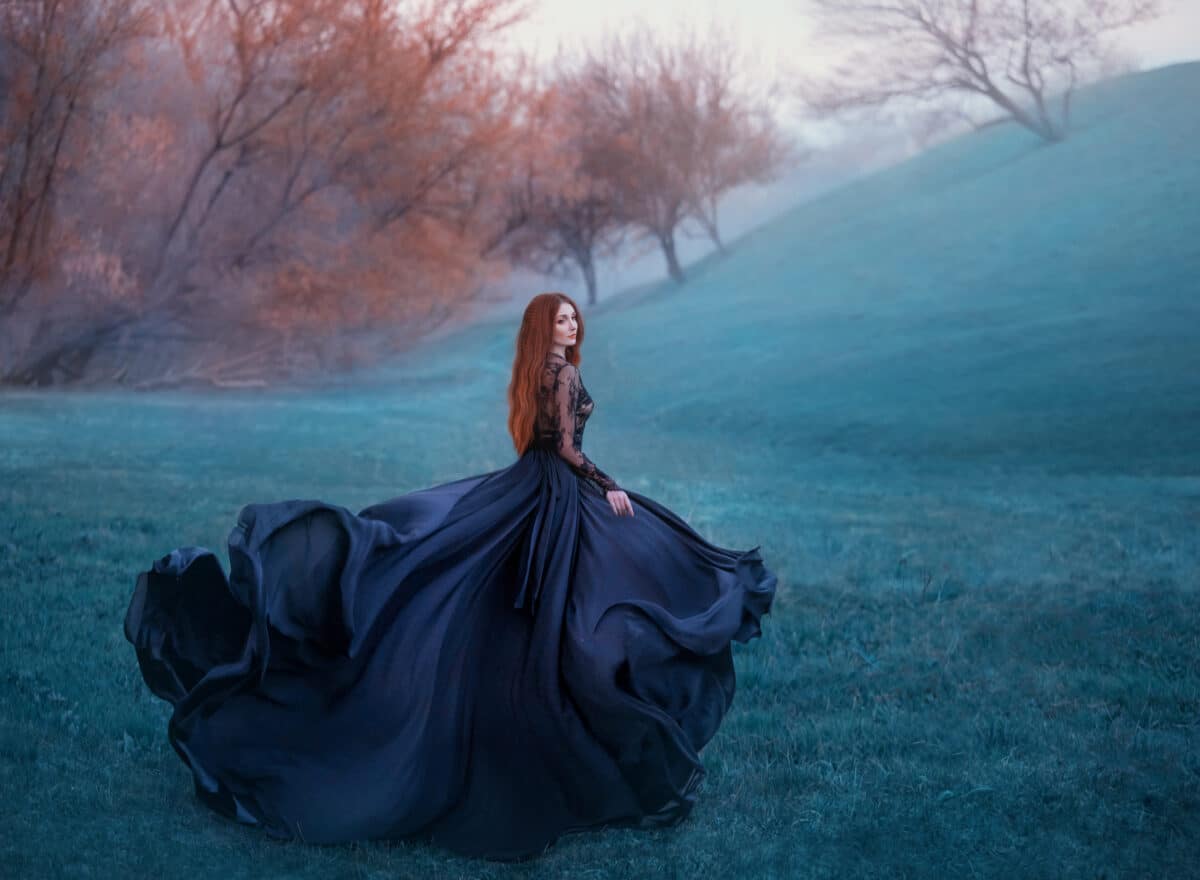charming lady with red hair on green hill, scraping widow in long black flying dress and lace blouse turns into flower, young sorceress looking for herbs in autumn cold forest, creative colors