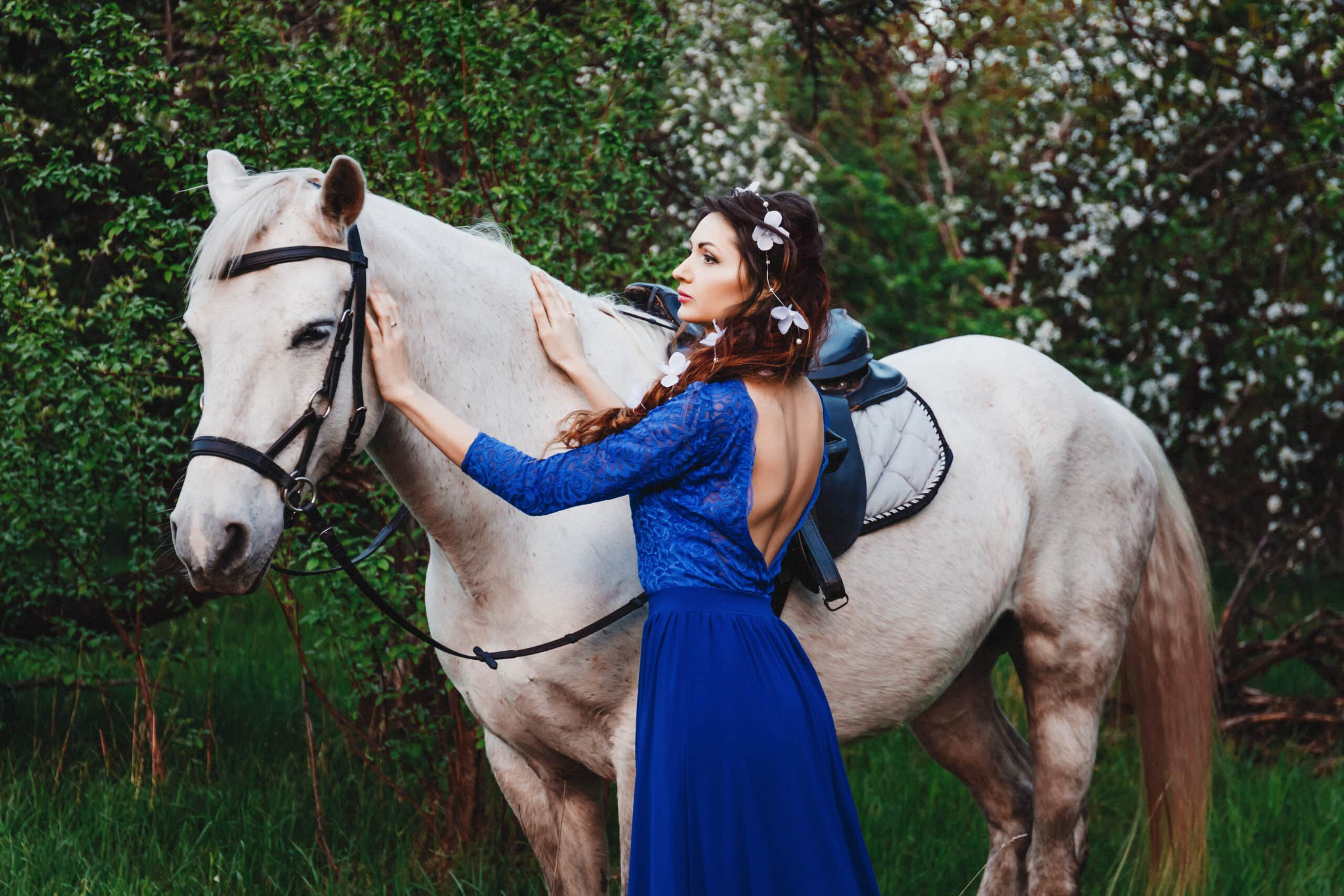 beautiful brunette woman in blue dress standing next to a white horse in spring.