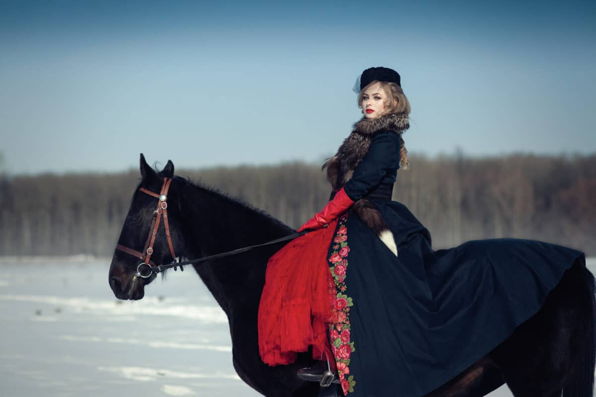 a blonde lady in a long black dress riding on a dark horse in winter