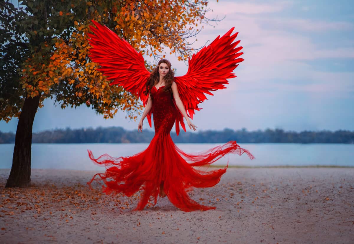 Young beautiful fantasy woman fallen angel lying in air near a tree with orange leaves. Creative red costume, huge artificial bird wings and elegant dress. magic autumn foliage. Photo of levitation.