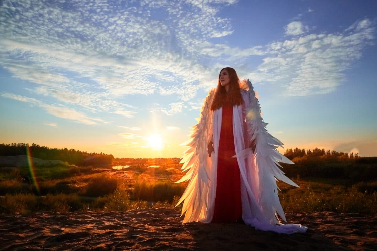 young woman dressed in red with white wings standing on the sand at sunset with the view of the light blue sky with feathery clouds