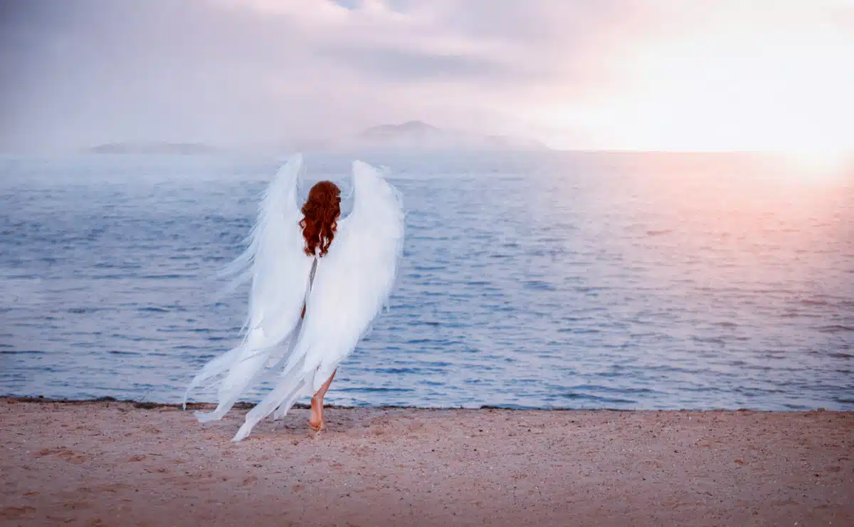 Young beautiful woman fallen angel stands on the sea beach enjoy nature. A costume huge artificial bird white wings. Silhouettes of a mysterious red-haired girl at sunset. Bright sunlight of sunset