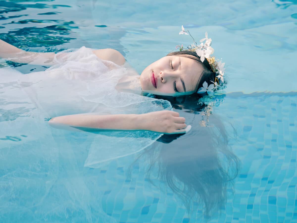 Beautiful fairy in a beautiful white wedding dress and garlands of flowers, floating in swimming pool with eyes closed, beautiful Chinese girl in water in sunny day.
