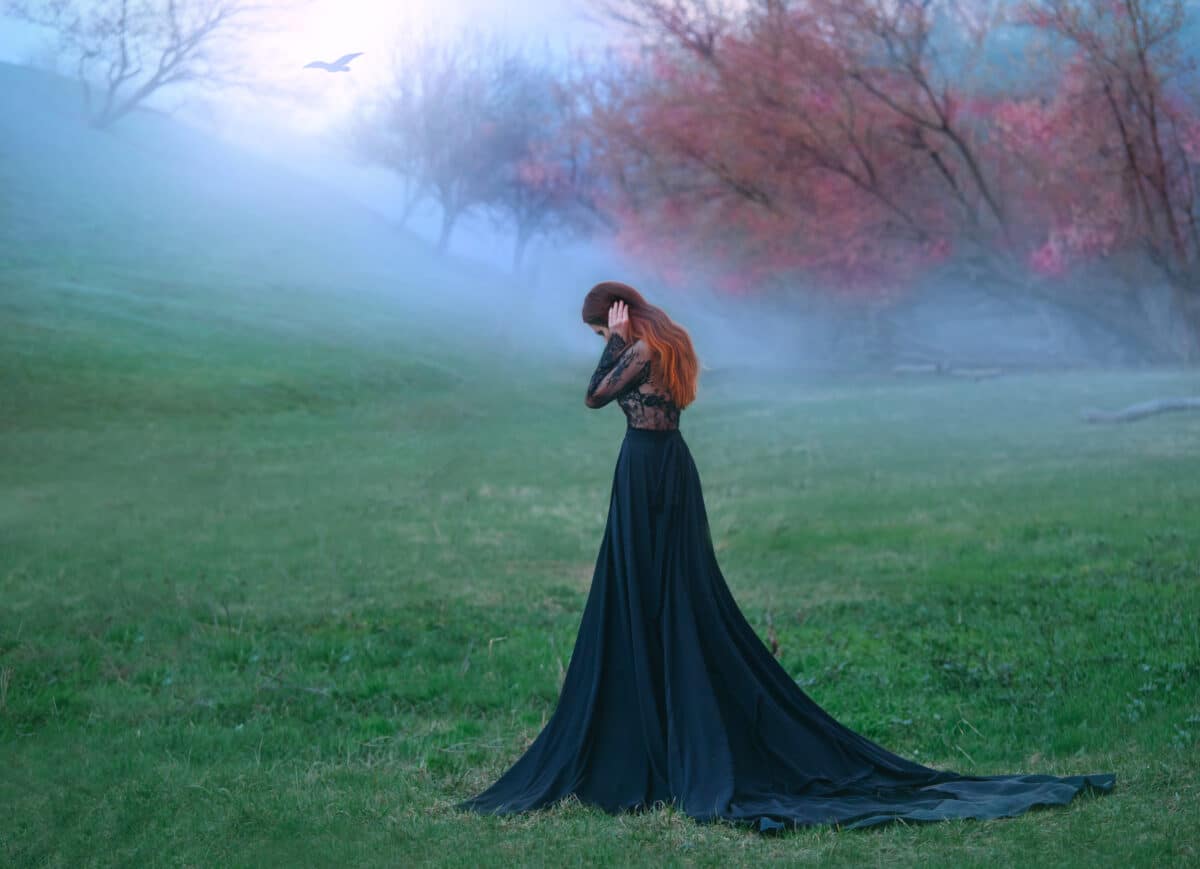 dark witch made terrible mistake, sad lady in long black dress with long tail and lace sleeves, girl with bright red orange hair pressed hands to head, autumn landscape, mysterious white haze
