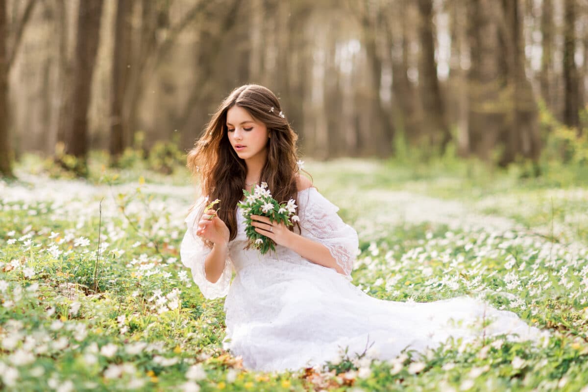 young beautiful woman in a white dress collects primroses. A girl in the spring forest. A bouquet of white anemones in her hands