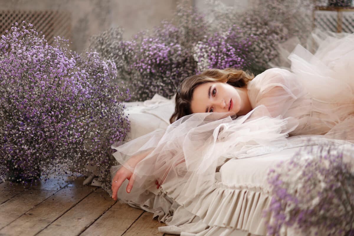 a young beautiful woman in tender transparent dress laying on bed among purple flowers