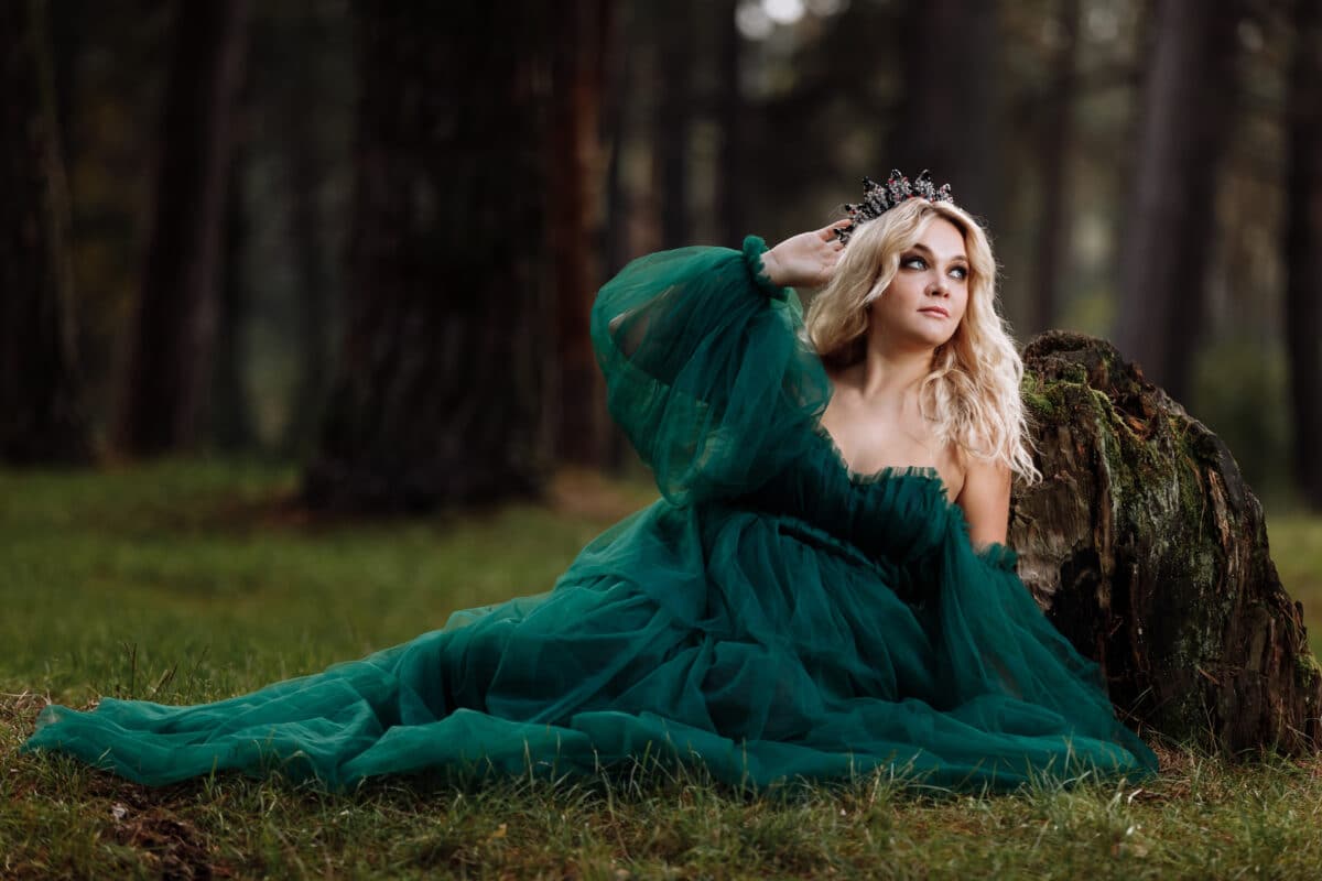 A beautiful blonde young woman in a long green dress and a diadem on her head in the forest. girl sitting near the old stump. Solar glare. Fantasy. fairy tale