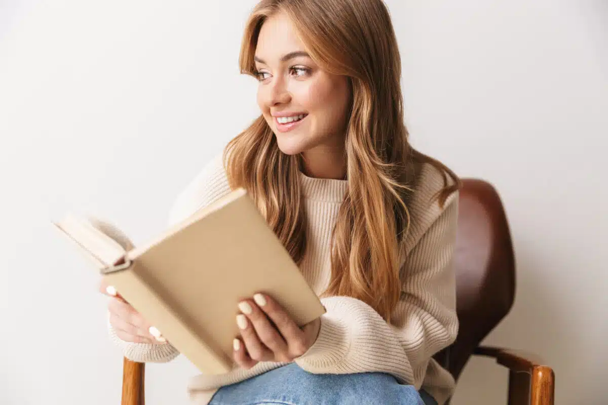 Image of happy young woman smiling and reading book while sitting