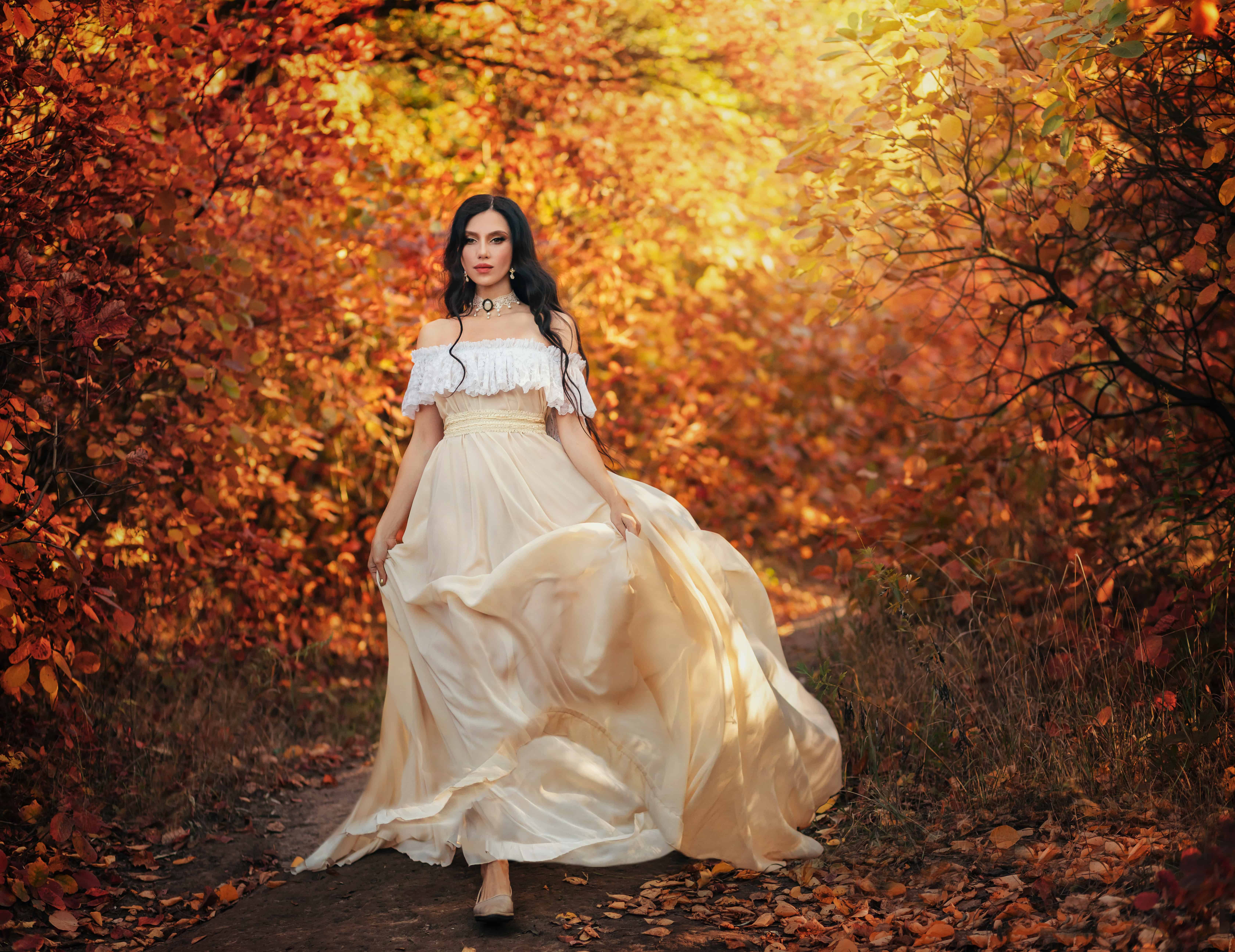 Art photo fantasy woman queen walking in gothic autumn forest, white vintage style dress. Girl princess beauty face long wavy hair, elegant sexy bare open shoulders. Red orange yellow color dark tree.