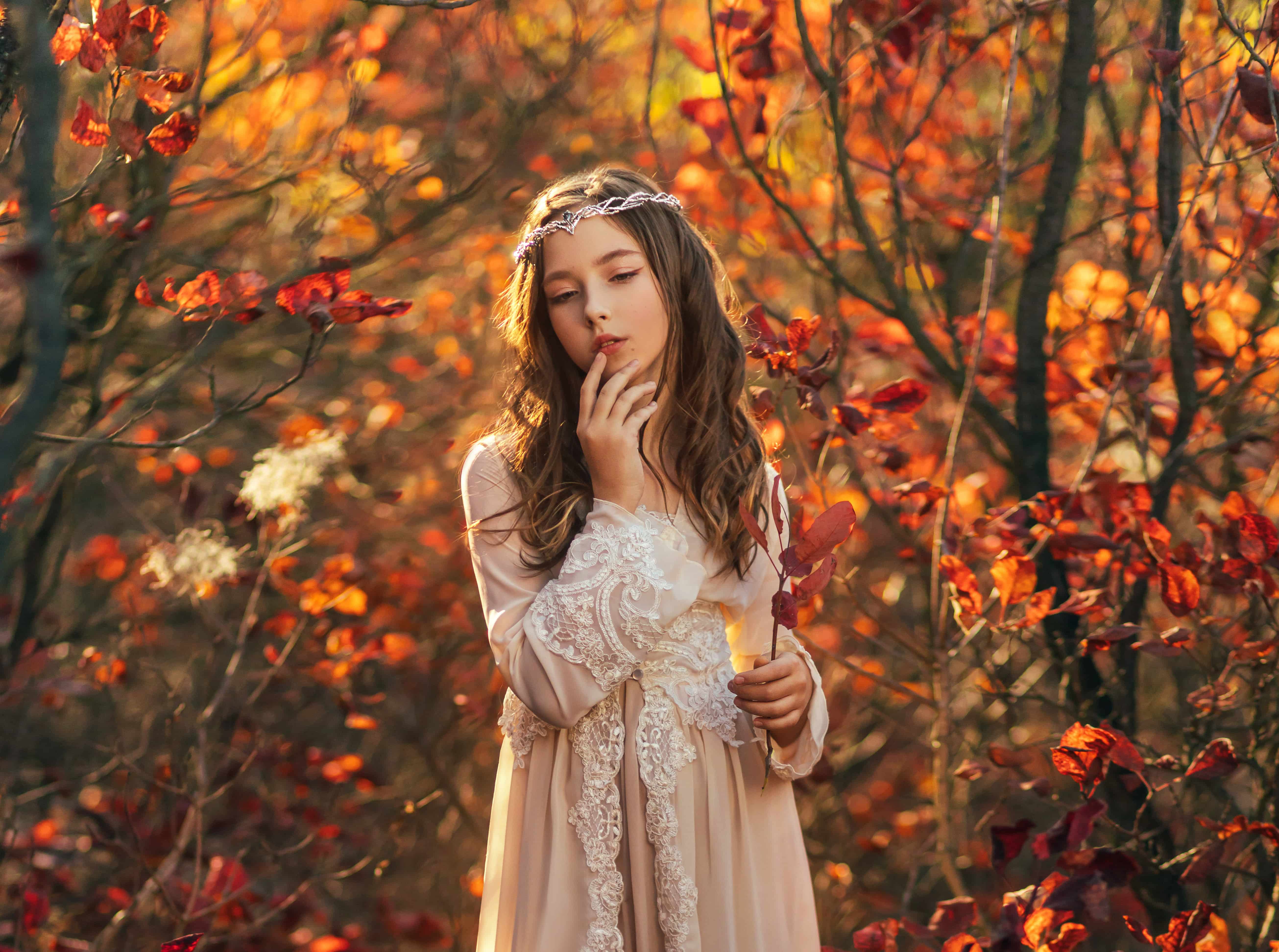 Fantasy portrait teenage princess girl walking in forest, blond flowing hair cute face. White vintage dress, silver crown tiara diadem on head. Autumn nature red orange leaves trees. Elf nymph woman