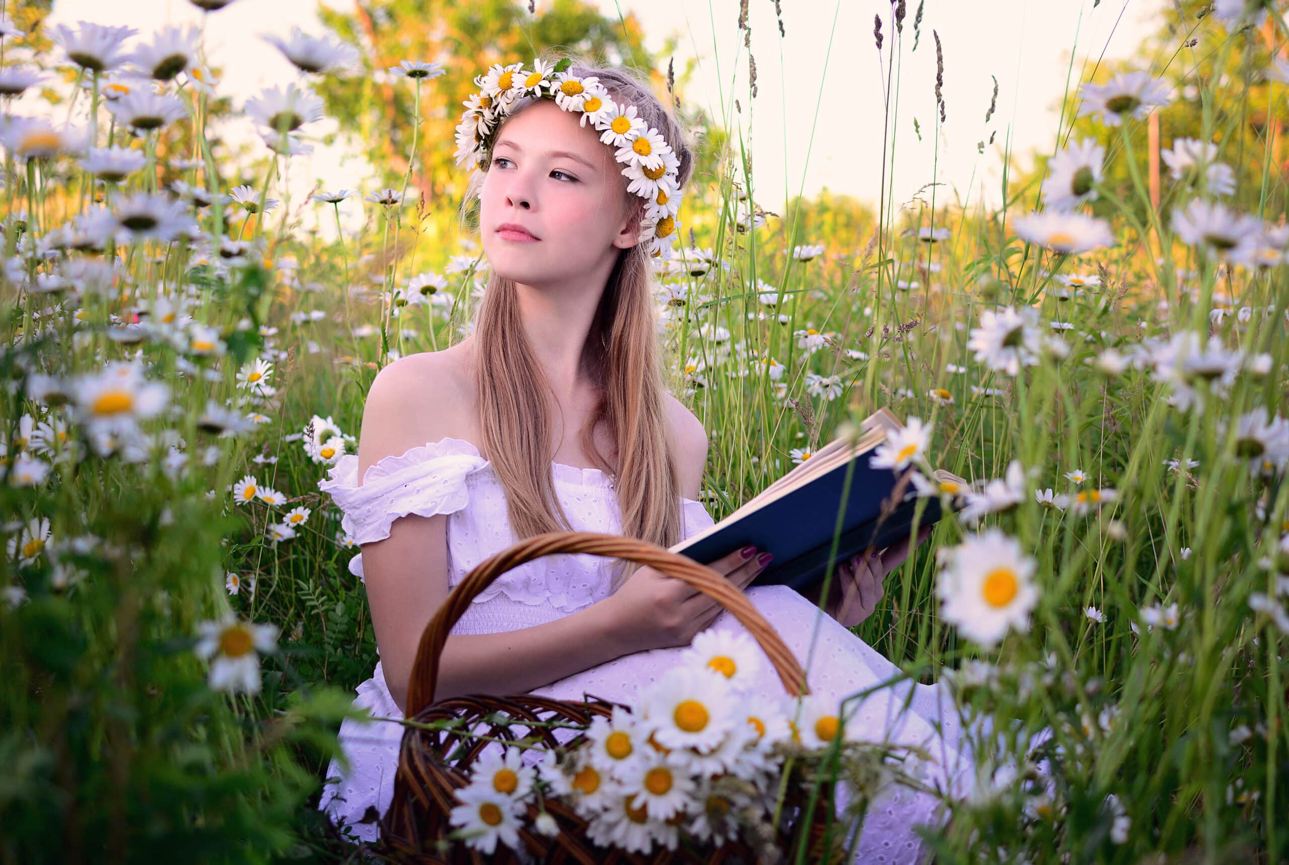 Young slim girl with a wreath on her head in a white dress sitting in a field of daisies with a book