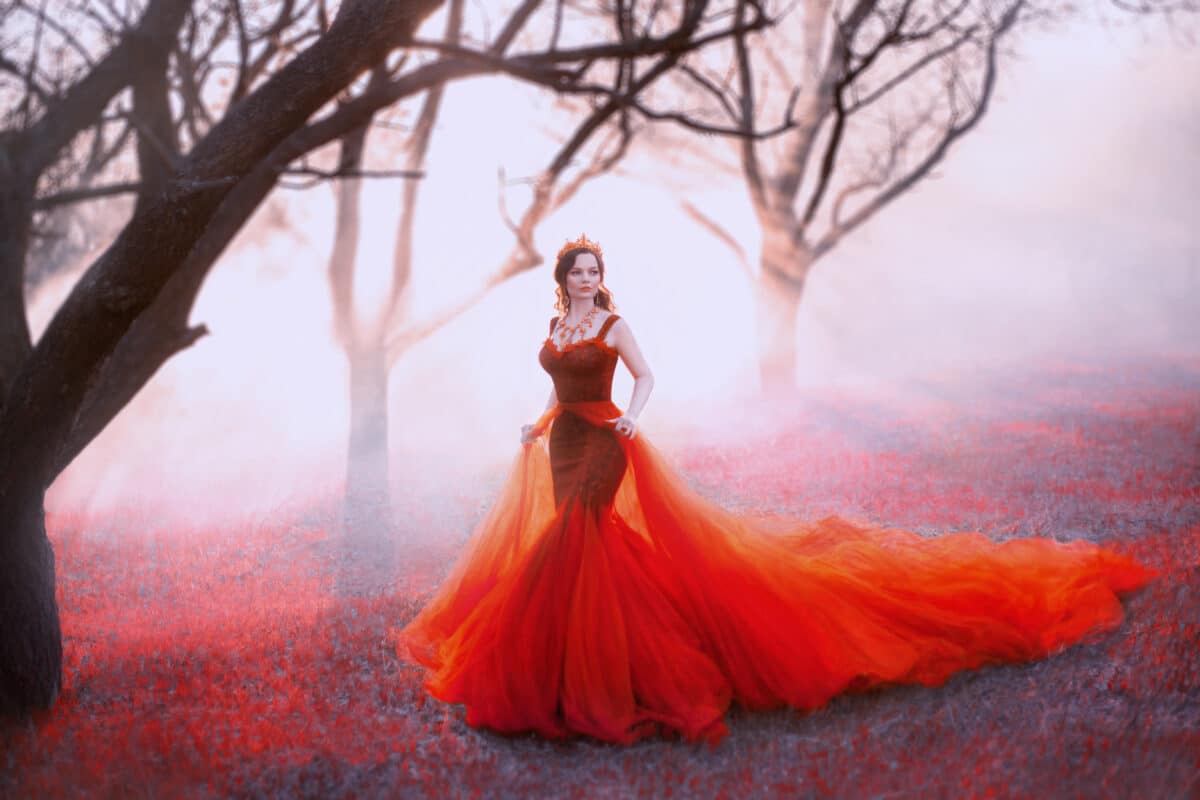 queen in long red dress with magnificent lush train, woman walks alone through scarlet autumn forest, gold crown and necklace on light body, royal charm and majesty, sun rays make through bare trees