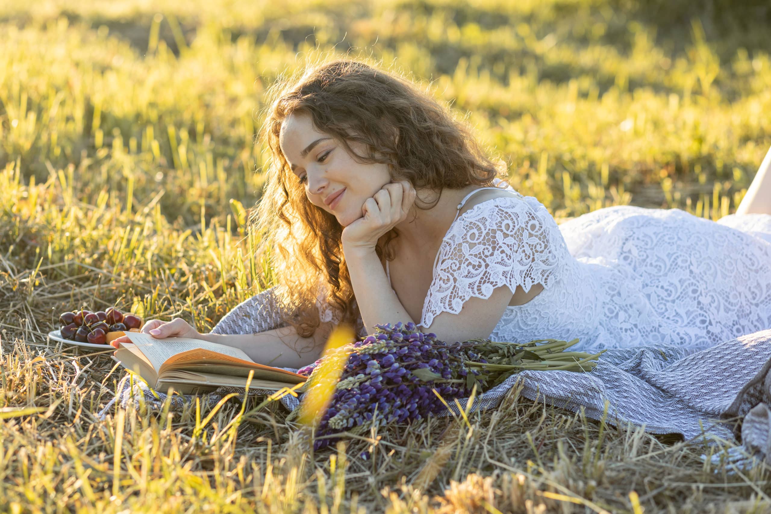 Beautiful young woman in a white dress, straw hat, picnic basket reading a book on a meadow.