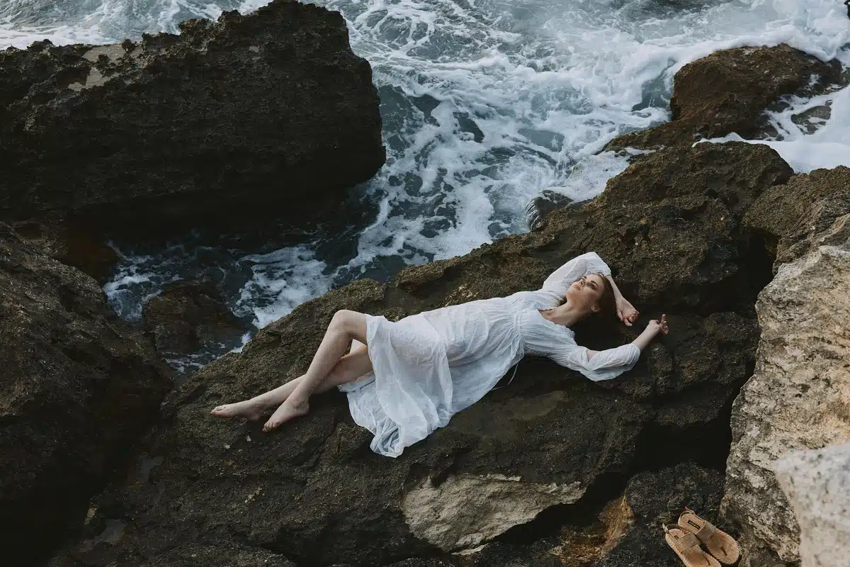 beautiful woman in long white dress wet hair lying on a rocky cliff by the frothy ocean