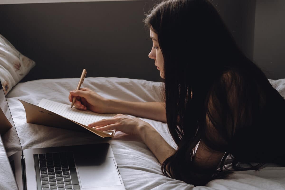 Young woman lies on bed with laptop and notebook and takes notes. Distance education, remote work