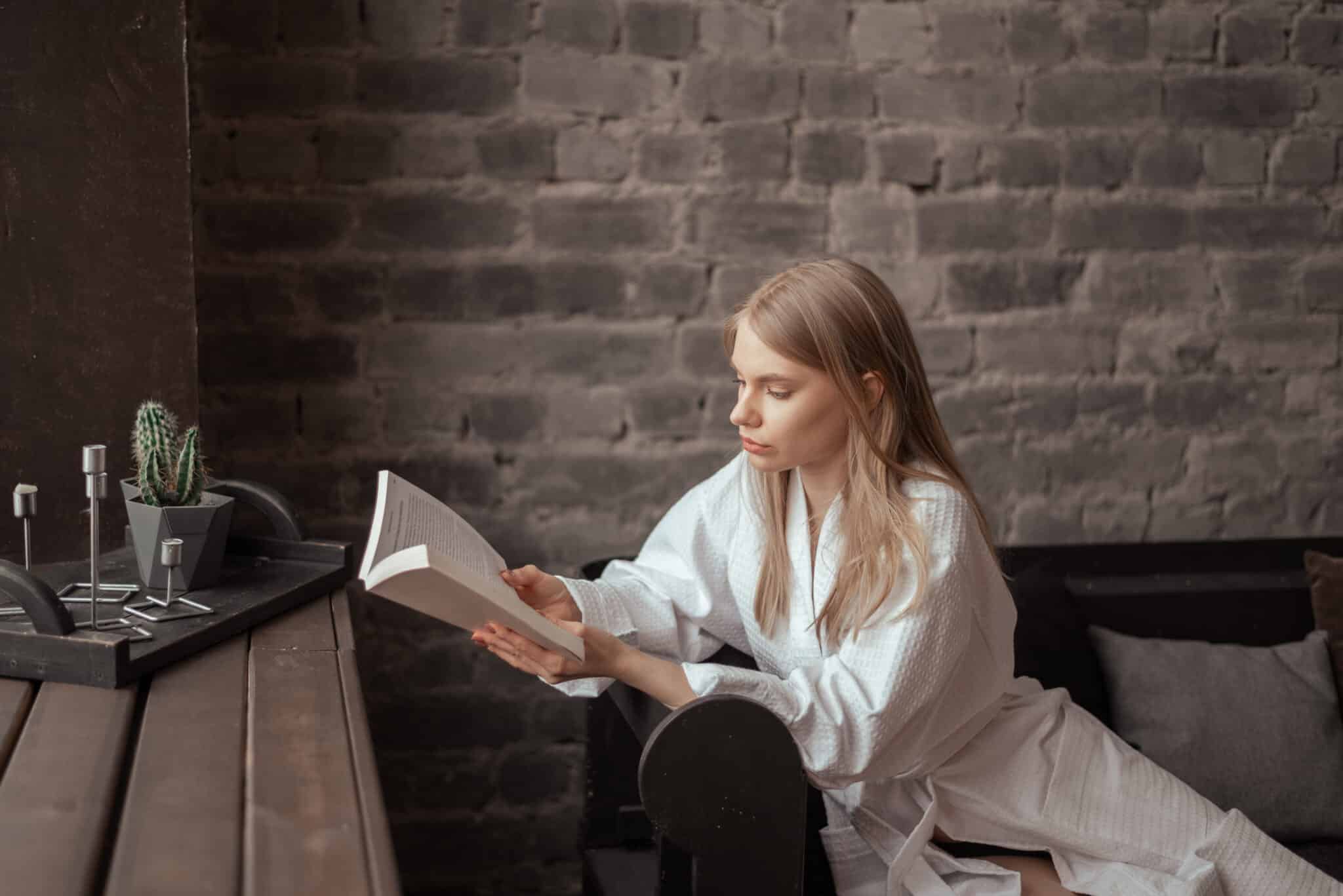 Young blonde beautiful woman in the white bathrobe sitting next to the window on the couch and reading the paper book.