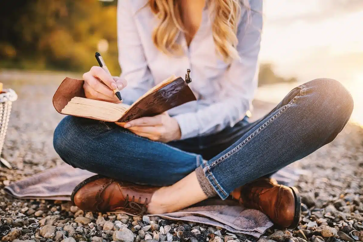 Woman in casual clothing sitting in lotus position, writing on her notebook outdoors.
