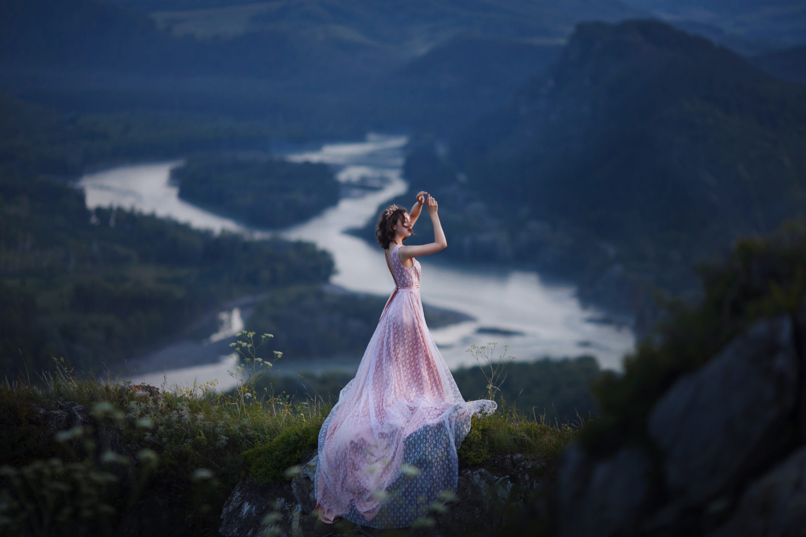 A girl in a pink dress with a long train is dancing on the top of a mountain.