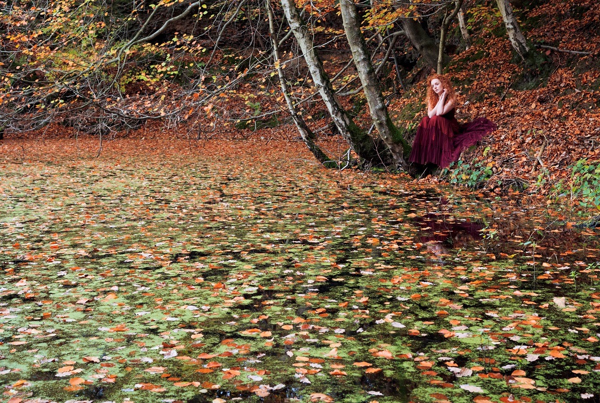 Beautiful woman in red corset and skirt with long red hair in the woods by the lake filled with fallen leaves..