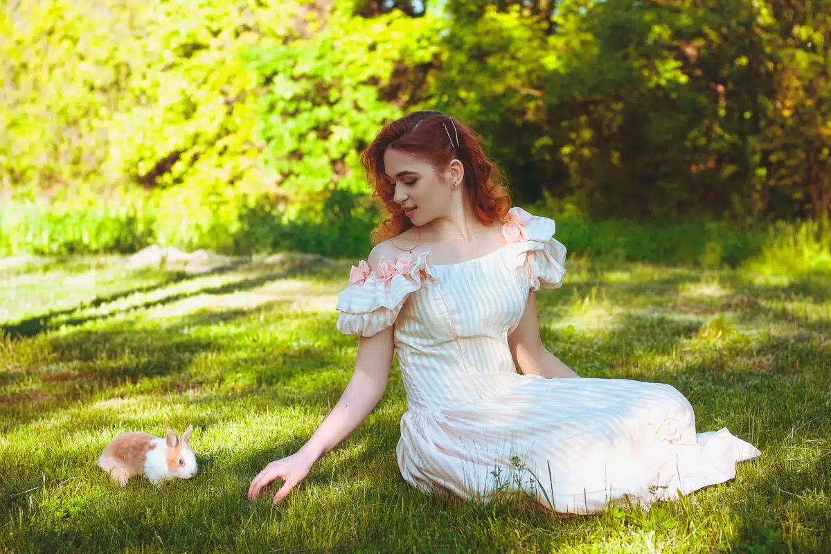 Young redhead woman in long dress playing with real cute fluffy bunny. Easter Day in green spring park. Female spring, summer fashion concept.