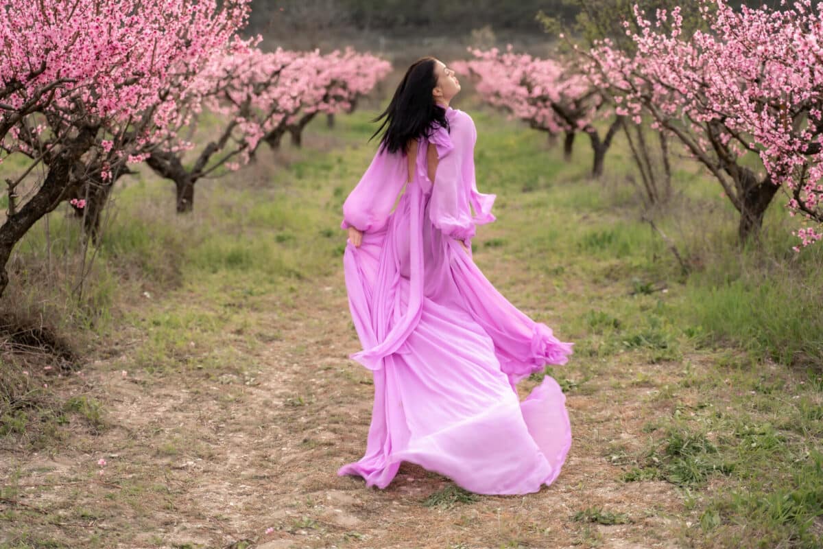 a woman in a long pink dress walks a blooming peach orchard