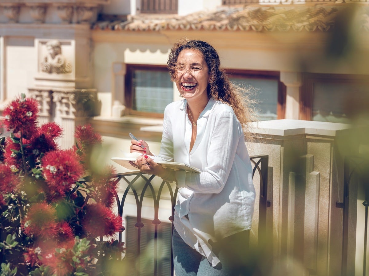 Cheerful woman writing notes in diary and laughing in balcony