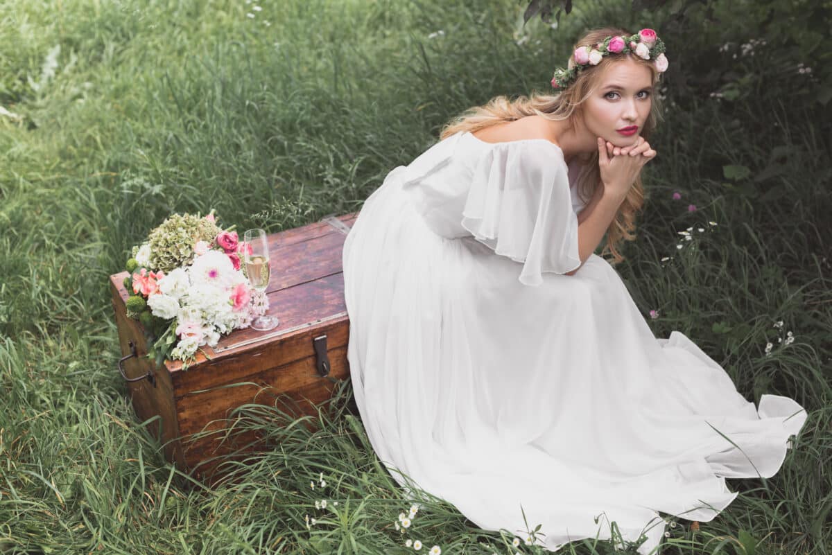 beautiful young woman in white with flower head wreath sitting on vintage chest in the garden