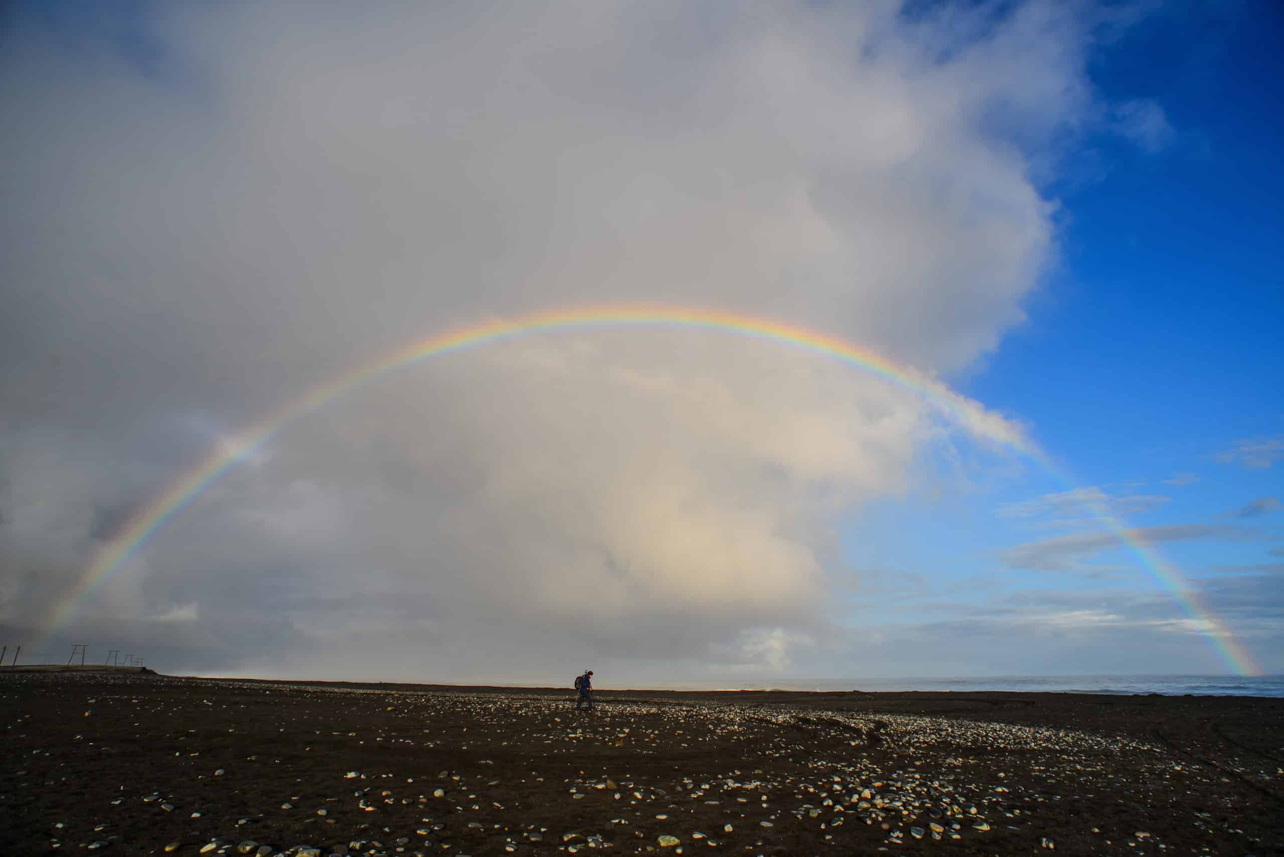 A man walking beneath a rainbow at the beach.