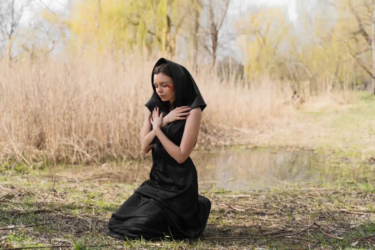 A young woman in a black mourning dress and headscarf kneeling on the ground, praying and crying with bloody tears