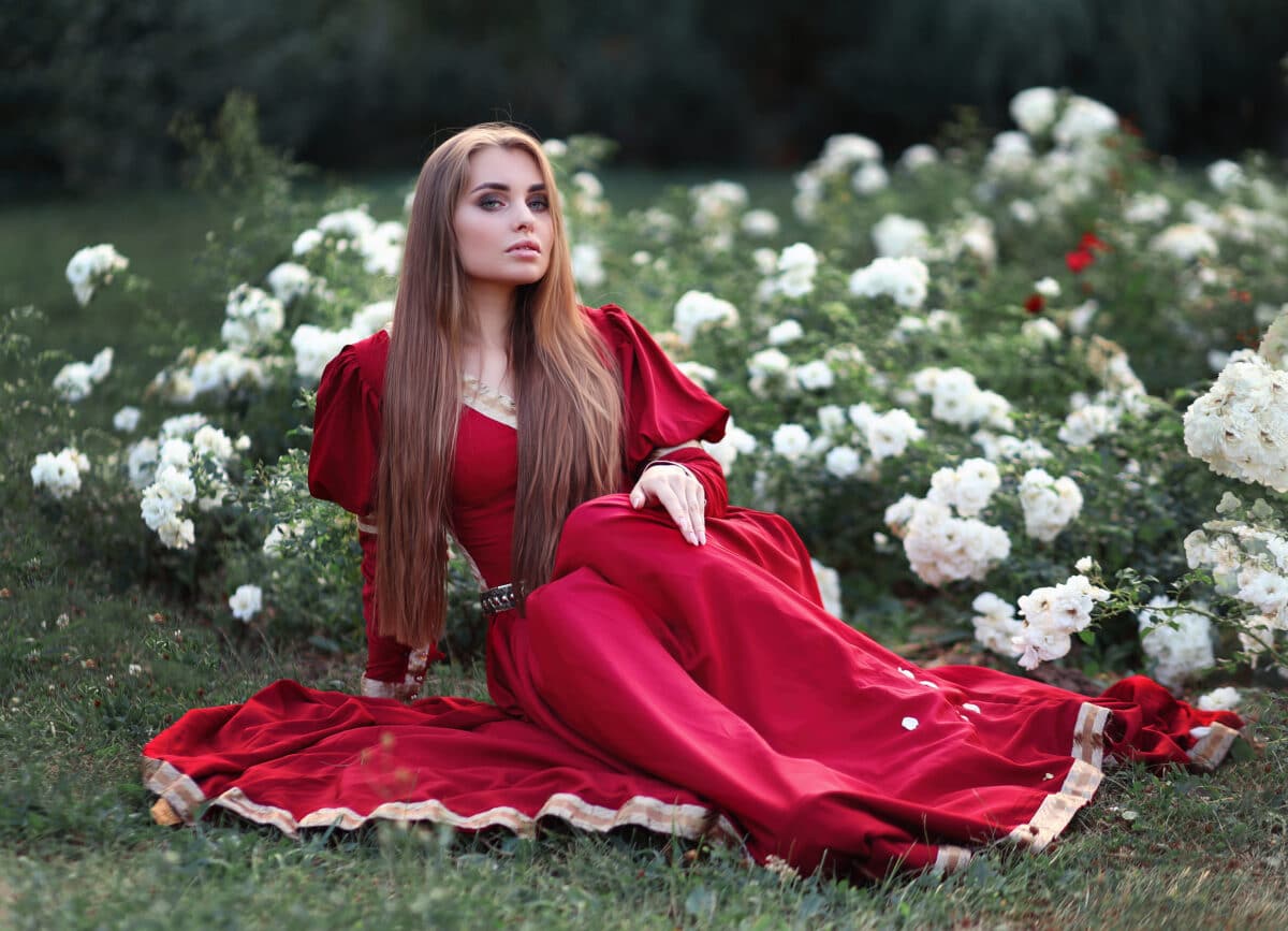 Beautiful young princess in a red dress sitting in the white rose garden